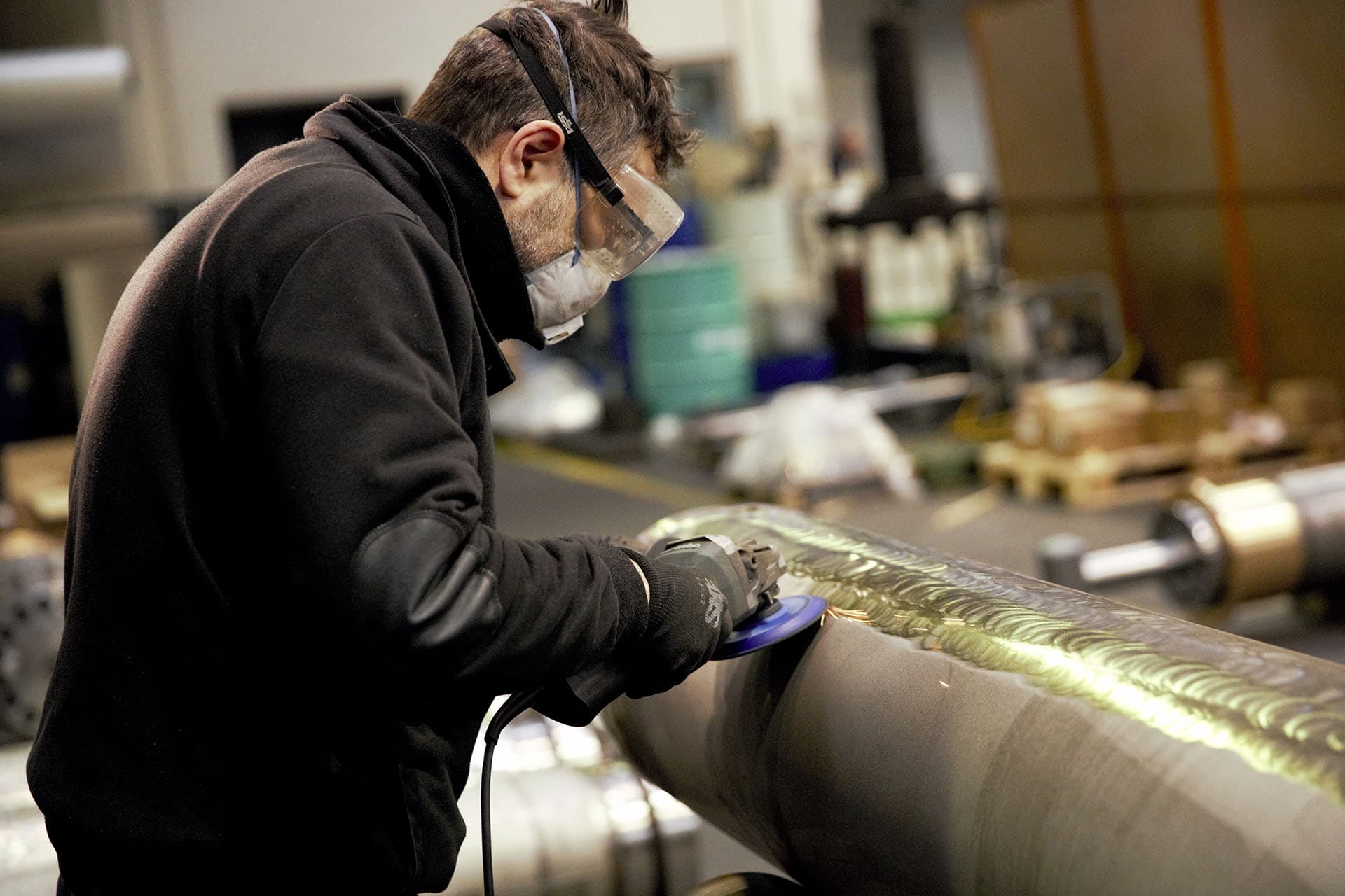 Opérateur avec lunettes et masque meulant la surface d'un grand vérin en acier