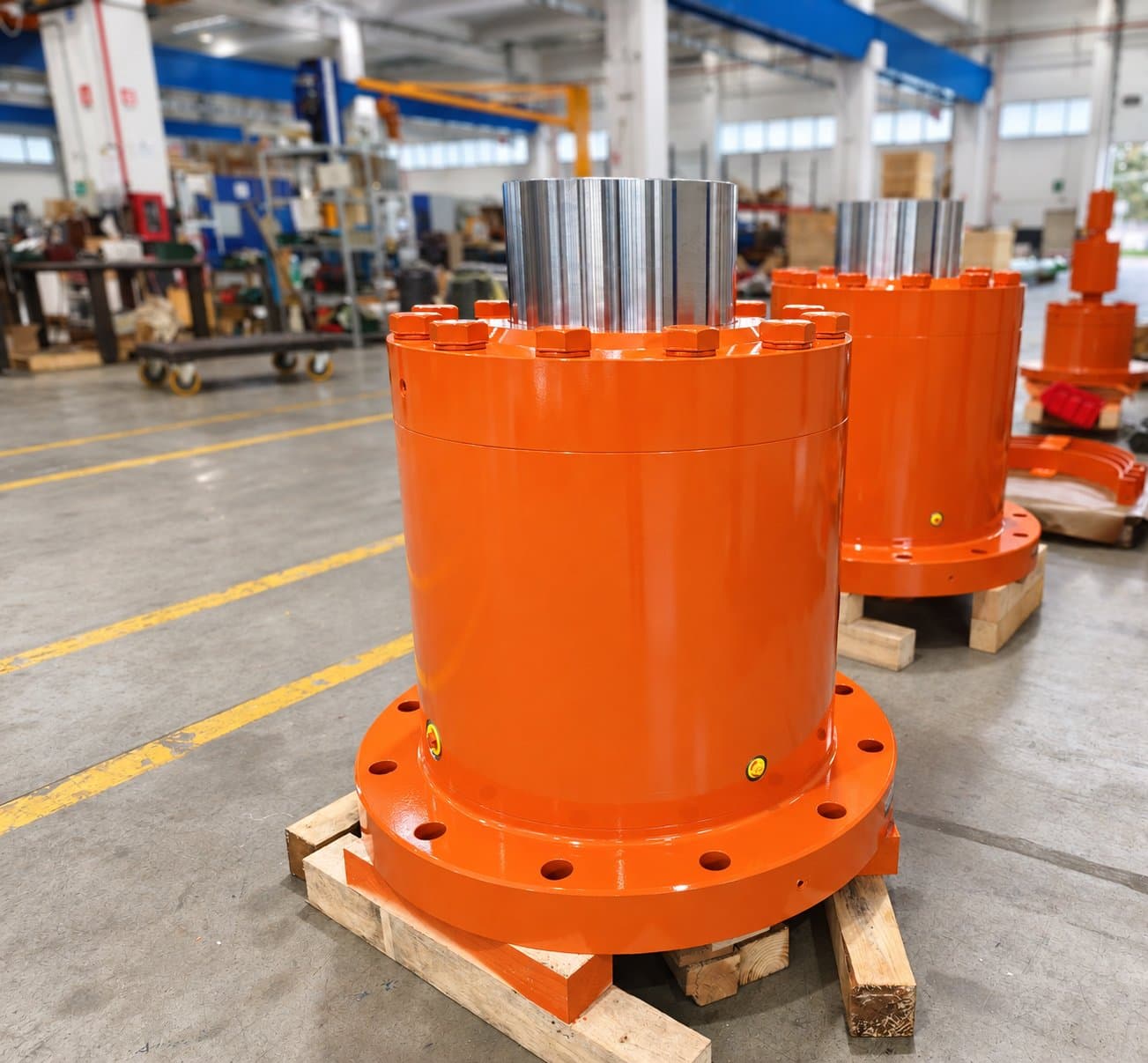 Two orange plunger cylinders with chromed rams and base flanges, on pallets in the workshop