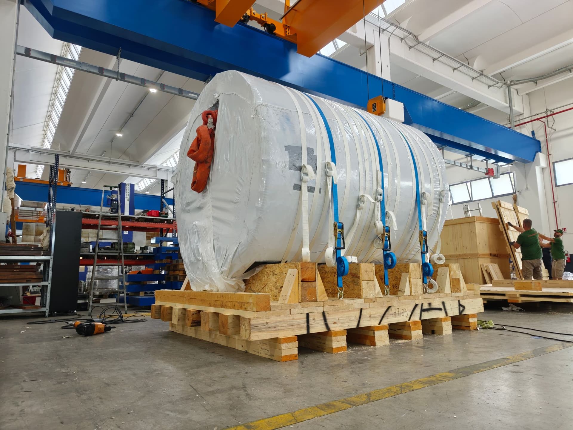 Wrapped cylinder strapped onto its wooden skid under the overhead crane, while operators assemble a shipping crate