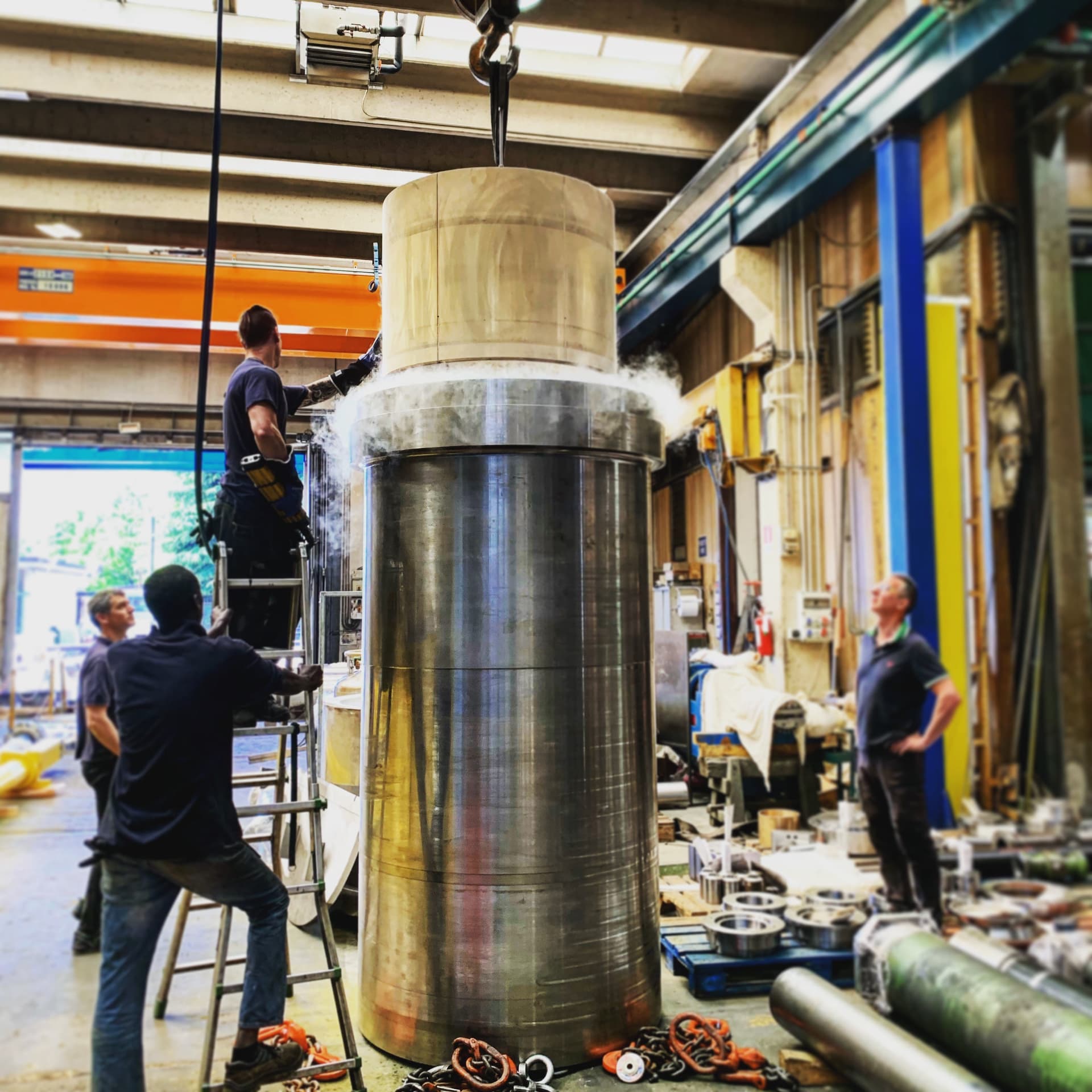 Workers around a large vertical steel cylinder being assembled in a workshop