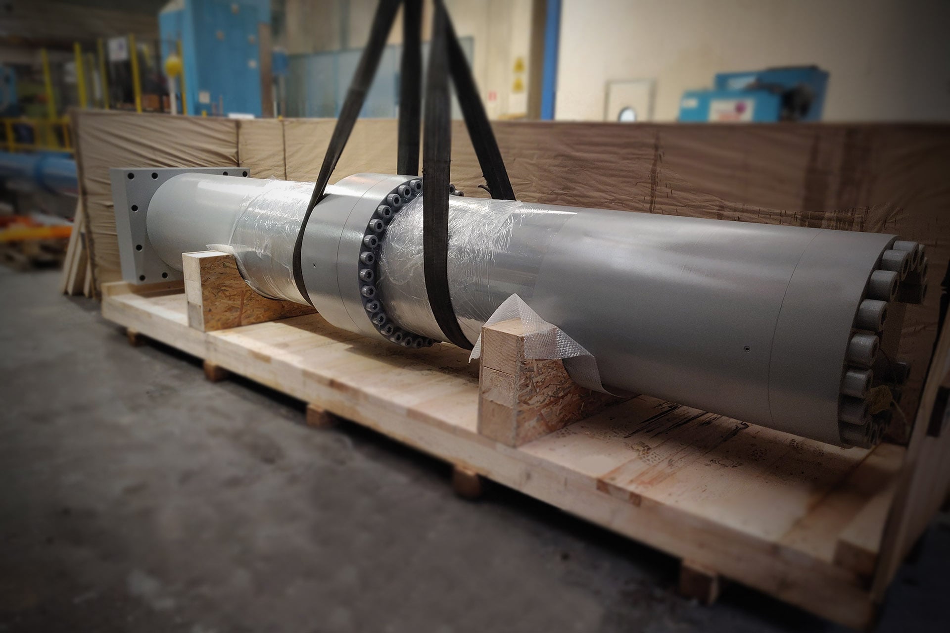 Grey hydraulic cylinder lifted by straps over a wooden base before packing